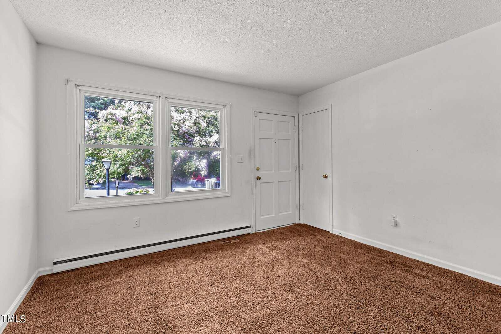 A small, empty room with white walls and a brown carpet. There are two windows showing a view of greenery outside, a white door, and a closed door leading to a closet. The room is well-lit with natural light.