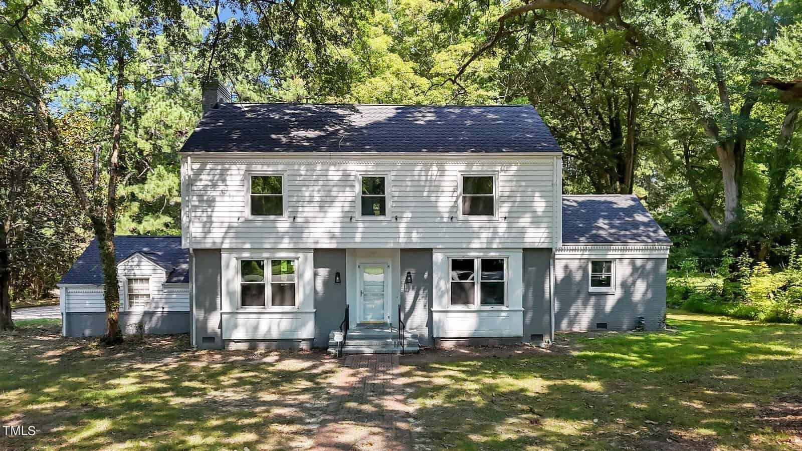 A two-story house with light gray siding and a dark gray roof, set amidst a lush, wooded area. The front of the house features multiple windows and a centered white door with steps leading up to it. Large trees provide shade over the lawn and the house.