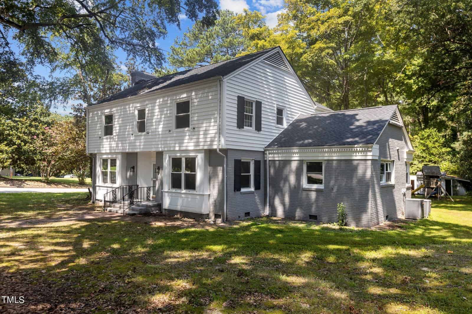 A two-story house with gray siding and white trim, nestled among large trees on a grassy lot. The home features several windows, shutters, a black railing, and a small porch. The sky is blue with a few clouds, and the area surrounding the house is shaded and green.