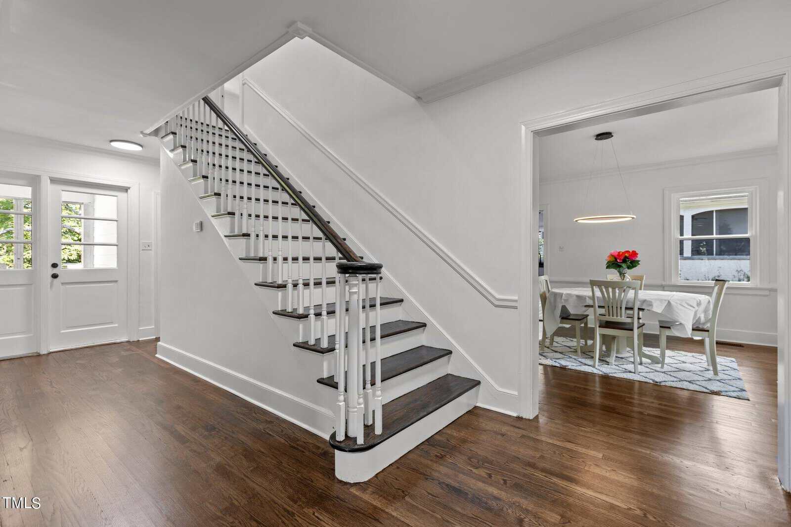 A bright, modern entryway featuring a wooden staircase with white risers and dark steps. Adjacent to the staircase is a dining area with a table, chairs, and a vase of red flowers, set on a patterned rug. The floor is hardwood, and walls are painted white.