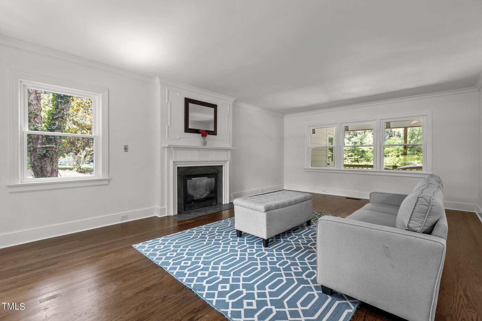 A minimalist living room features a gray couch and matching ottoman on a blue geometric rug. The room is bright with large windows and a fireplace with a mirror above it. Hardwood floors and white walls give the space a clean, modern look.