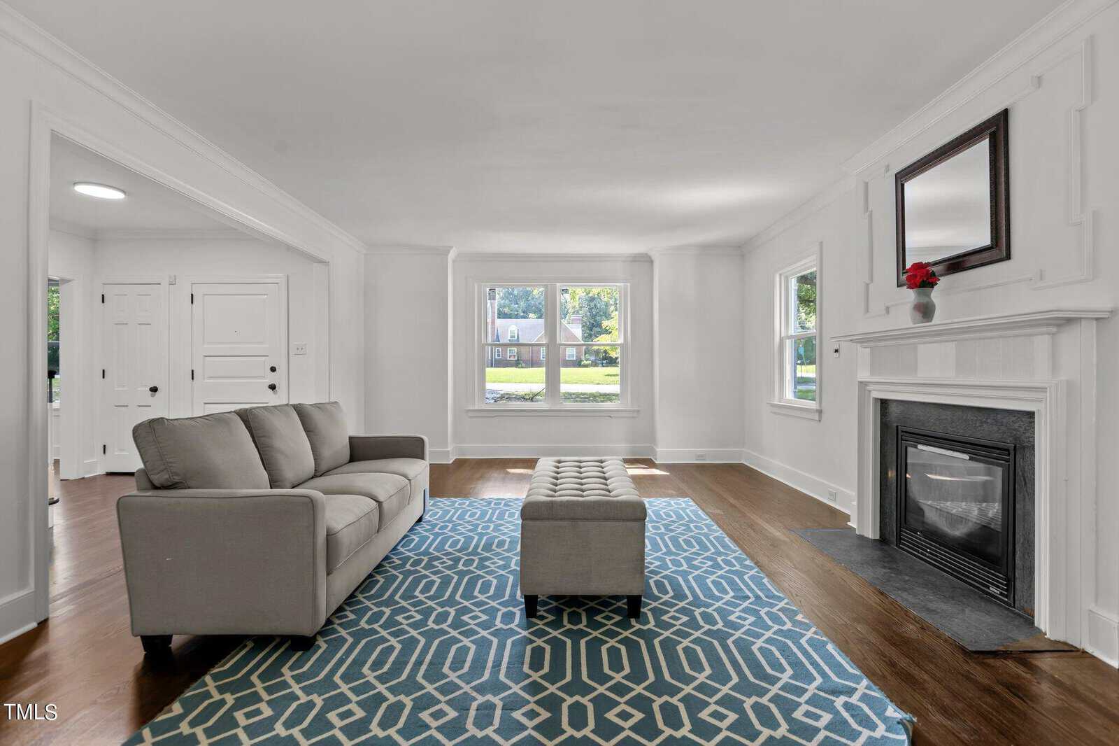 A bright living room with a gray sofa, upholstered ottoman, and a blue geometric rug. The room has large windows, a fireplace with a dark mantle and mirror above, and white walls. There's a glimpse of the adjacent room and the entryway with doors in the background.