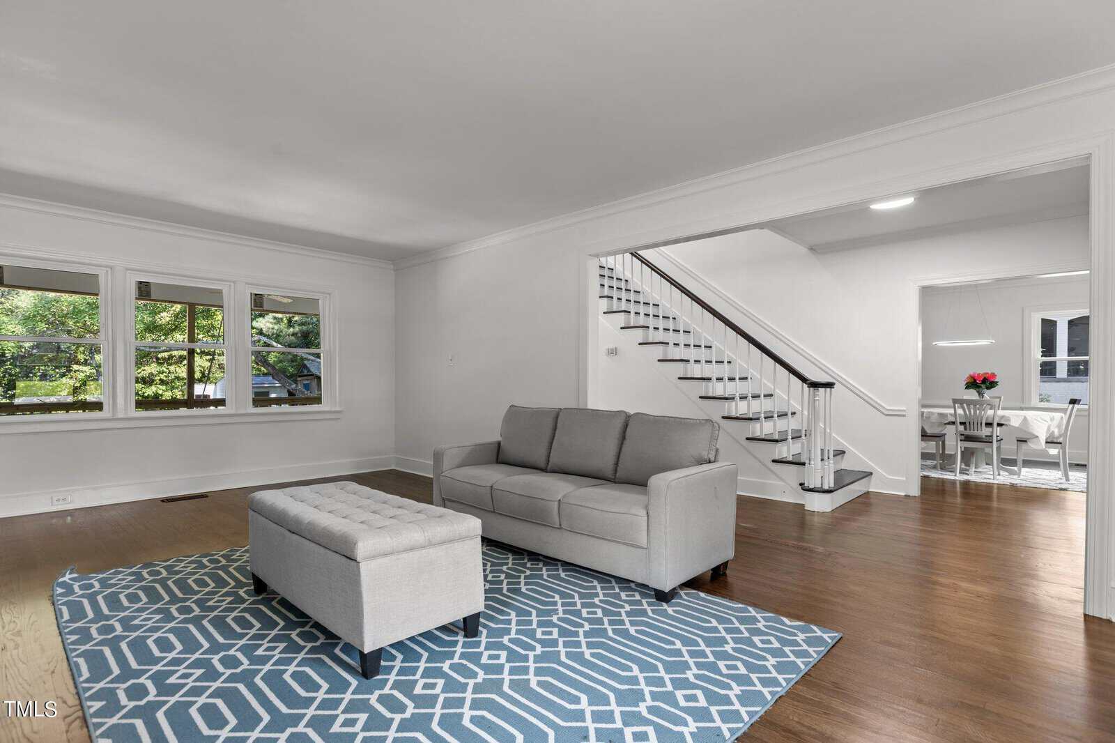 A spacious living room with white walls, large windows, and hardwood floors. It features a gray couch, a matching upholstered bench on a blue geometric-patterned rug, and a staircase leading to the upper floor. A dining area is visible in the background.
