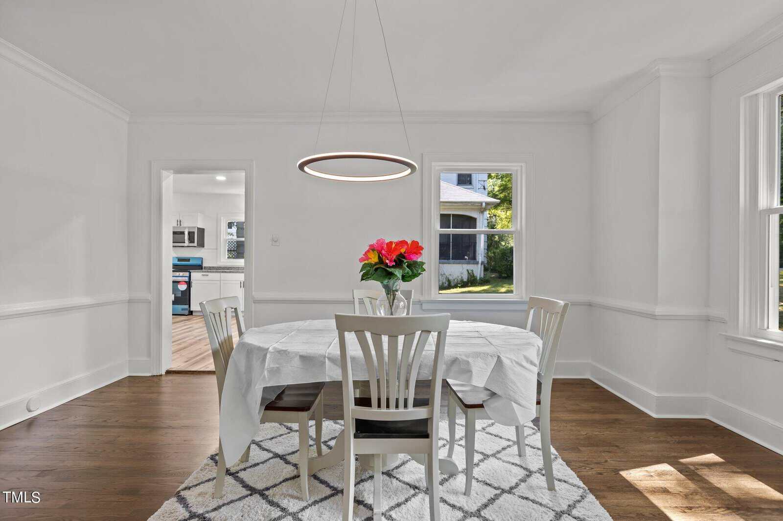 A bright dining room features a rectangular table covered with a white cloth, surrounded by four white chairs. A circular pendant light hangs above the table, which has a vase of vibrant red flowers. The room has white walls, large windows, and hardwood floors.