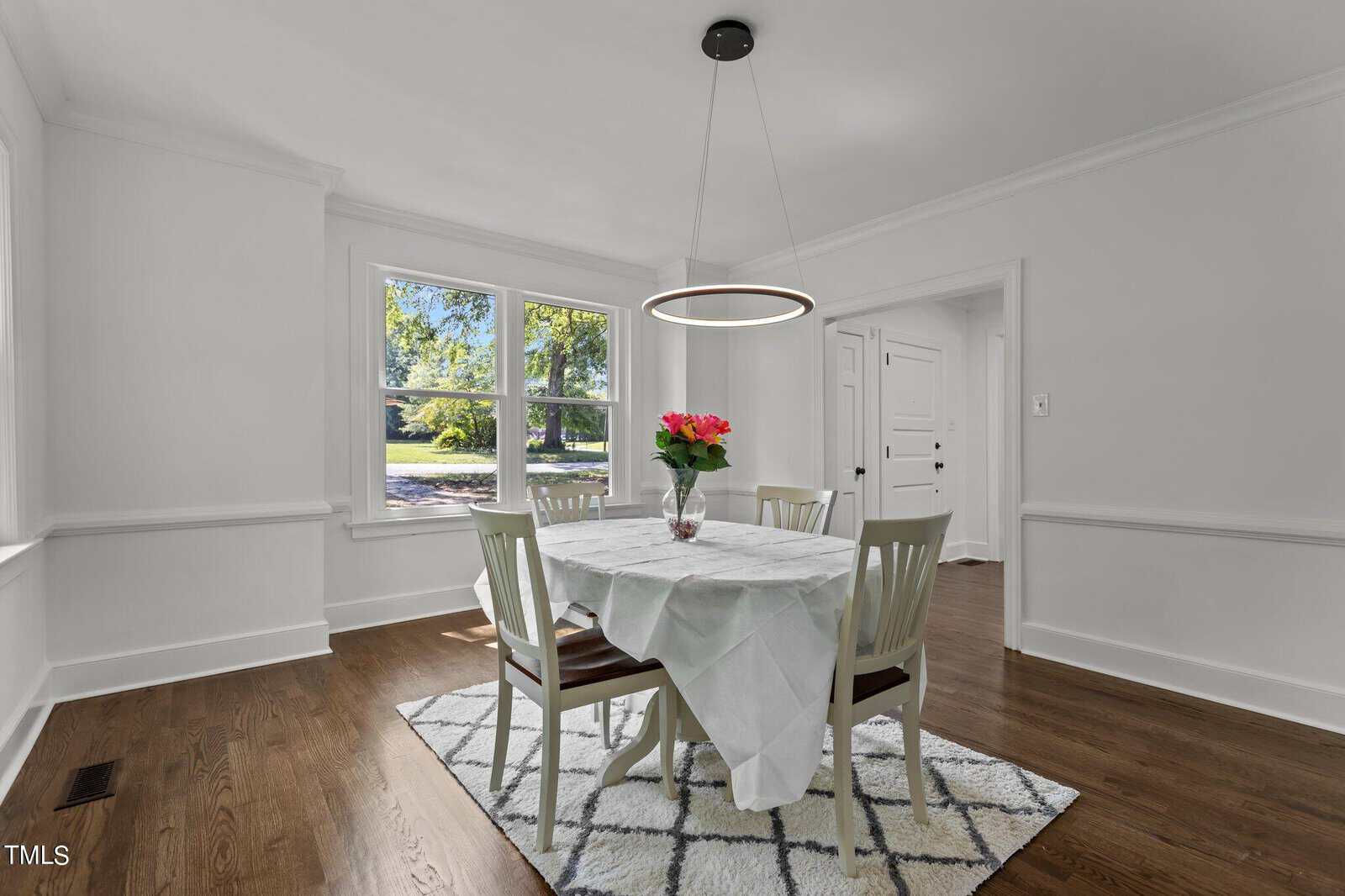 A bright dining room features a round table with a white tablecloth, surrounded by four white chairs. A vase of red and pink flowers sits on the table. The room has a modern light fixture, large windows with a view of greenery, white walls, and a hardwood floor.