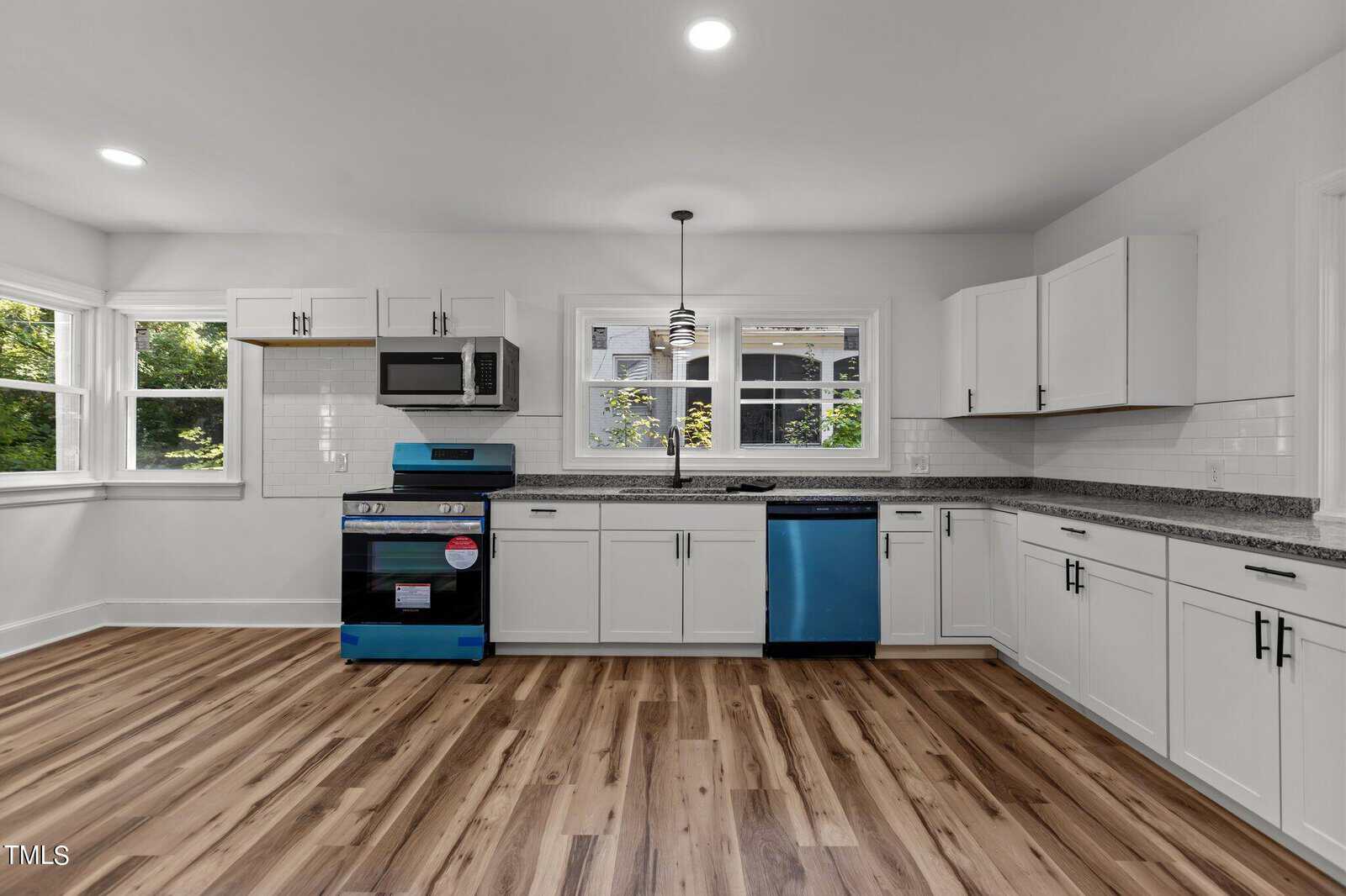 A modern kitchen with white cabinets, granite countertops, and stainless steel appliances. The dishwasher and oven are both wrapped in blue protective film. The floors are wooden, and there are large windows bringing in natural light.