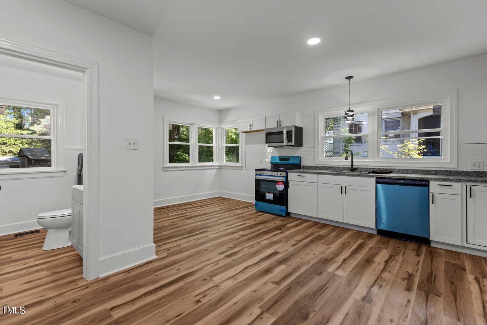 A modern kitchen with white cabinets, stainless steel appliances including a dishwasher and microwave, and a blue-protected oven. The kitchen has wooden floors and large windows allowing natural light. A door to the left opens into a small room with a toilet.