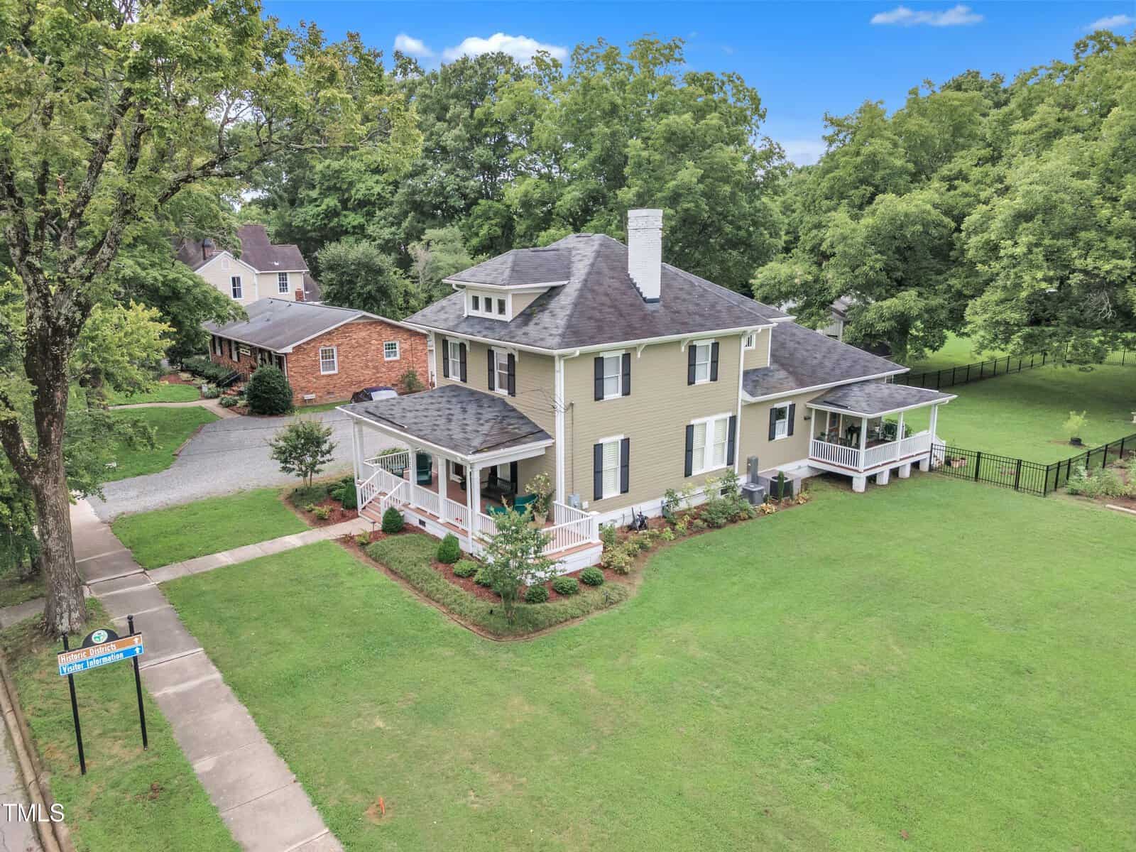 Aerial view of a spacious two-story house with a large front porch. It is surrounded by a well-maintained lawn and trees. A sidewalk leads to the front entrance and a neighboring brick house is visible in the background.