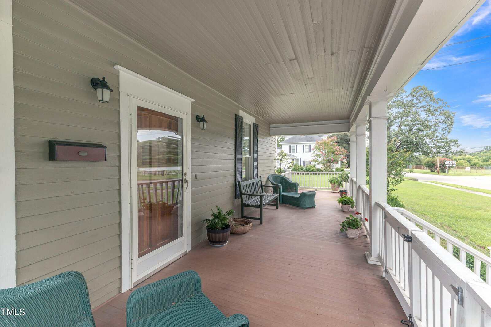 A covered porch with wooden flooring features green wicker chairs, potted plants, and a small table. The porch overlooks a grassy yard and street, with a view of neighboring houses and a clear blue sky in the background.