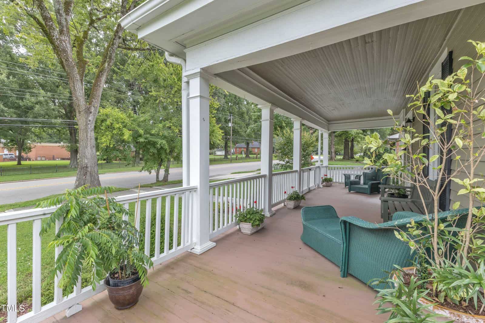 A spacious front porch with white railings and columns, overlooking a tree-lined street. Furnished with green wicker chairs, a small table, and potted plants, the porch offers a cozy, outdoor seating area.