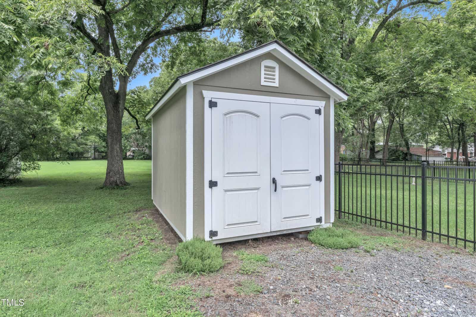 A small, beige garden shed with white double doors is situated on a gravel path next to a black metal fence. The shed is surrounded by green grass and trees, creating a serene outdoor setting.