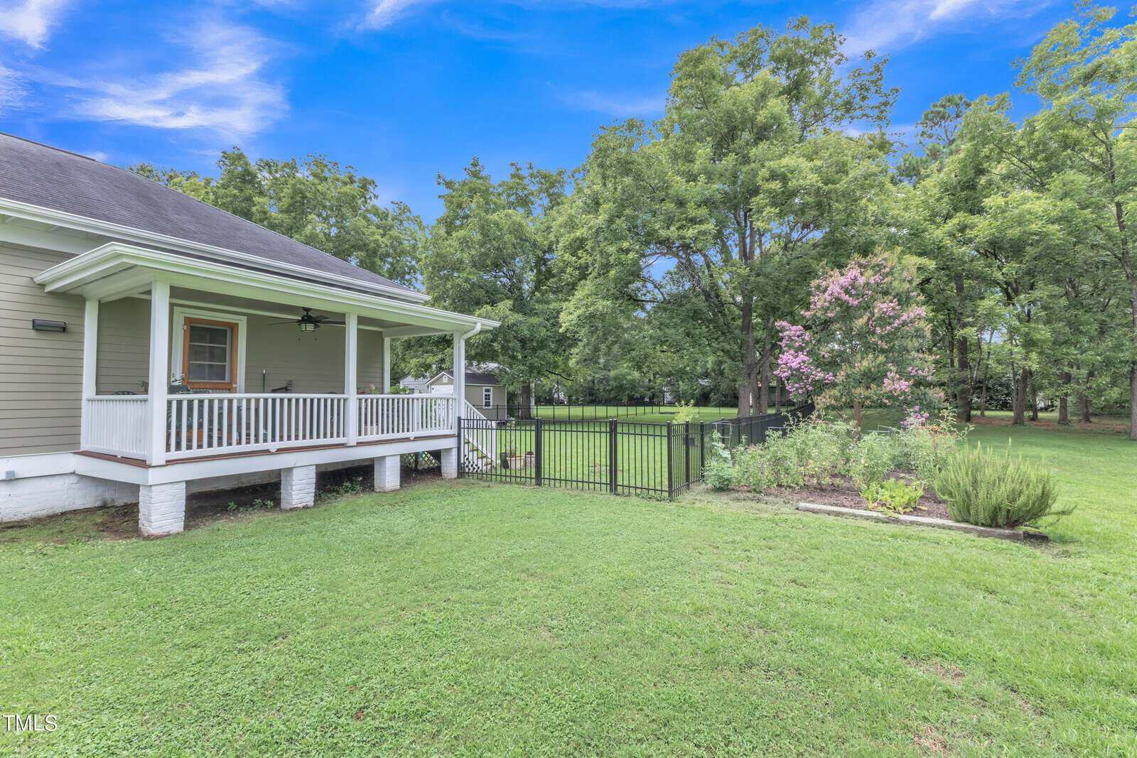 A peaceful backyard with a green lawn and mature trees. A wooden house has a porch, and a small flower garden with pink blooms is near a black fence. The sky is clear and blue.