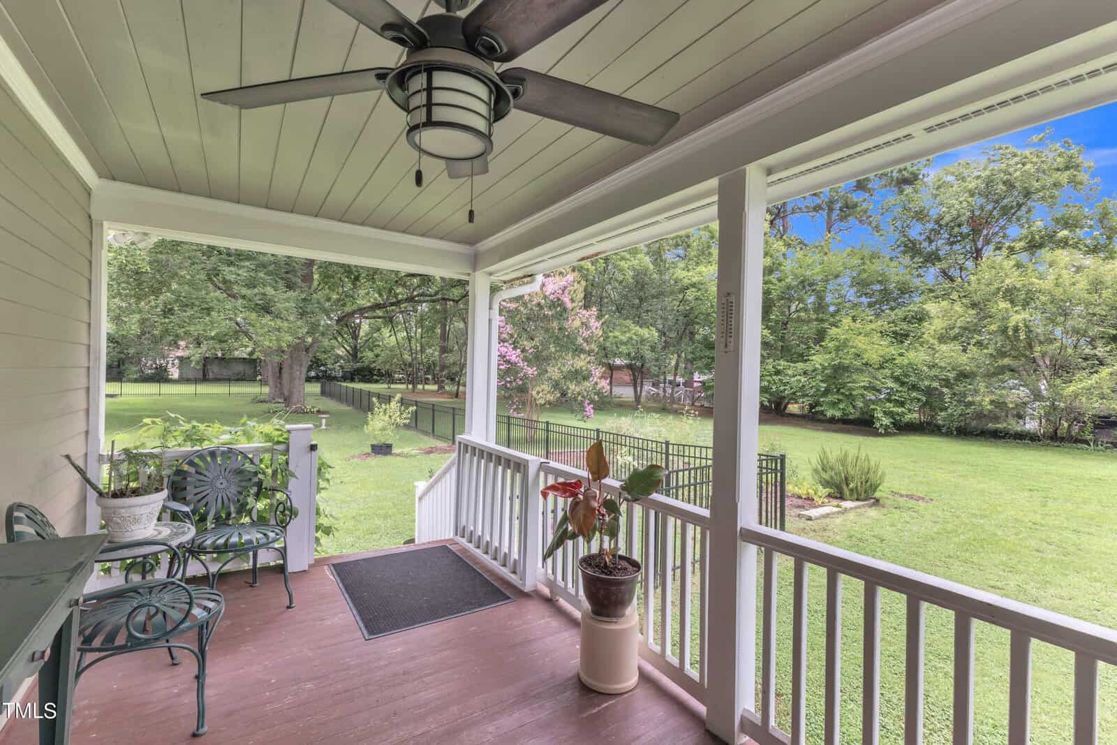 A cozy porch with a ceiling fan overlooks a lush, green backyard. There are two metal chairs and a small table on the wooden floor, next to a potted plant. Trees and a fence are visible in the background under a clear blue sky.