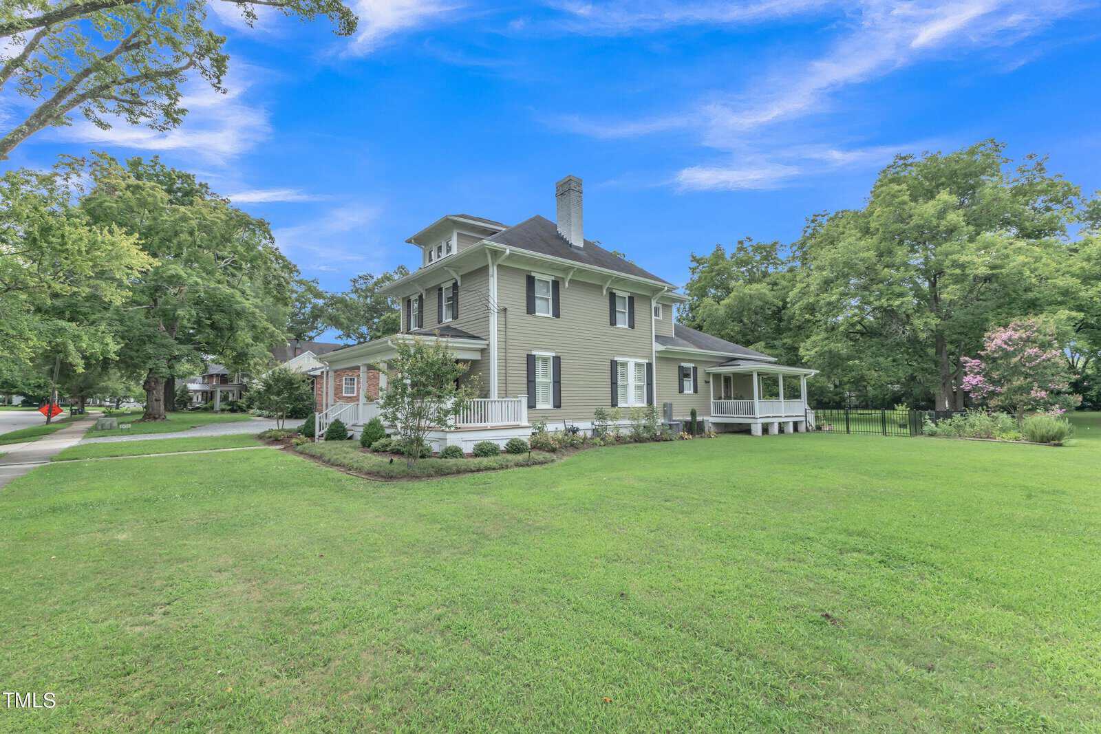 A large, two-story green house with white accents sits on a spacious, well-maintained lawn. It features a wraparound porch and is surrounded by trees and a bright blue sky.