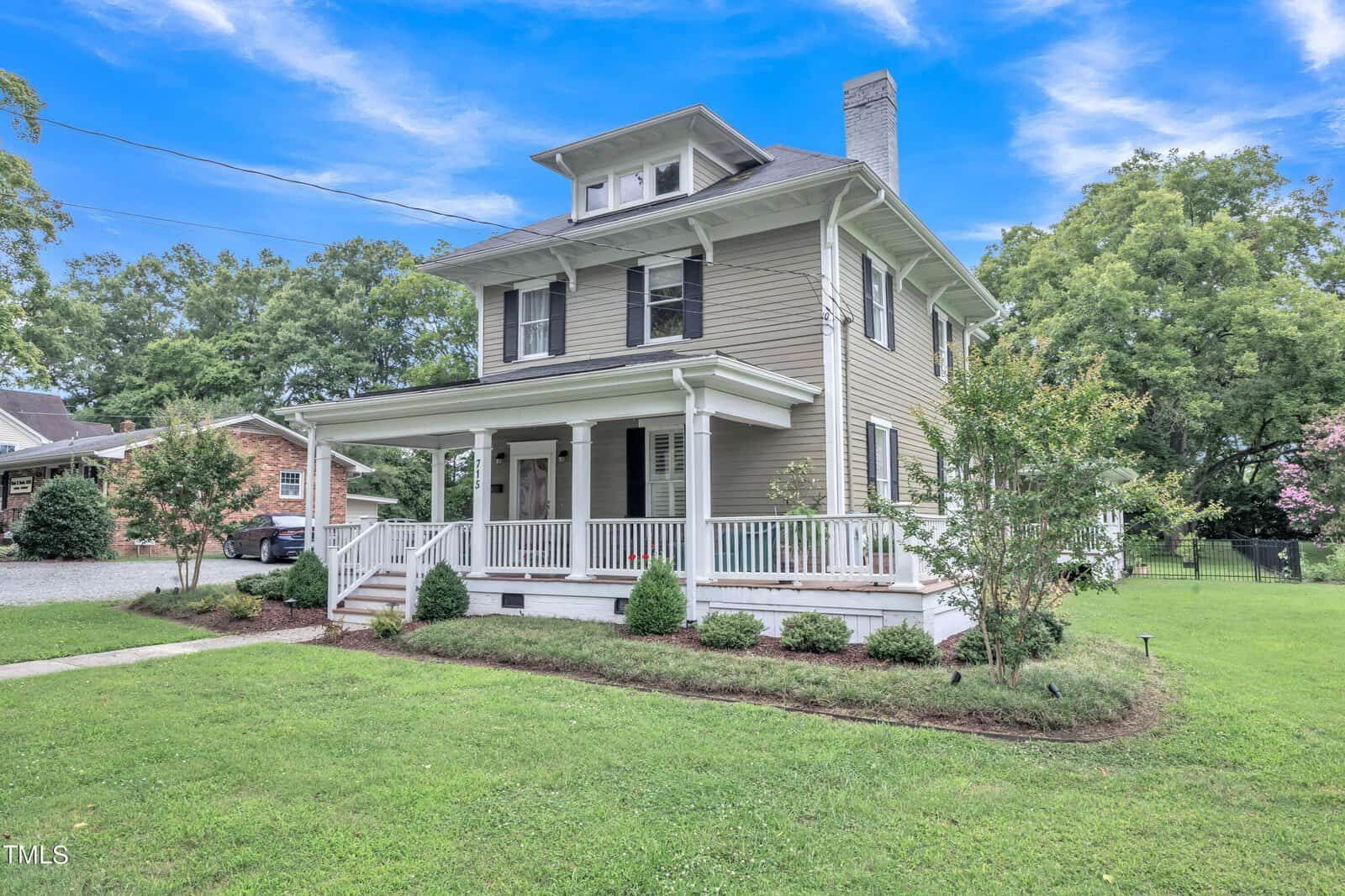 The charming two-story house at 715 College Street in Oxford features a wraparound porch and elegant gray siding. It's surrounded by a lush green lawn and trimmed bushes, while a gravel driveway leads to the small garage beside the home, all under a bright blue sky with scattered clouds.