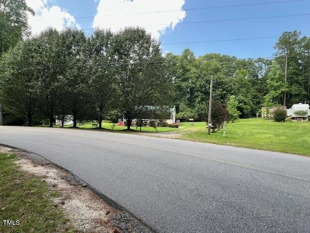 A two-lane road curves past a grassy area with trees, a mailbox, and a white house with a green roof. Power lines stretch above and an RV is parked on the right. The sky is partly cloudy.