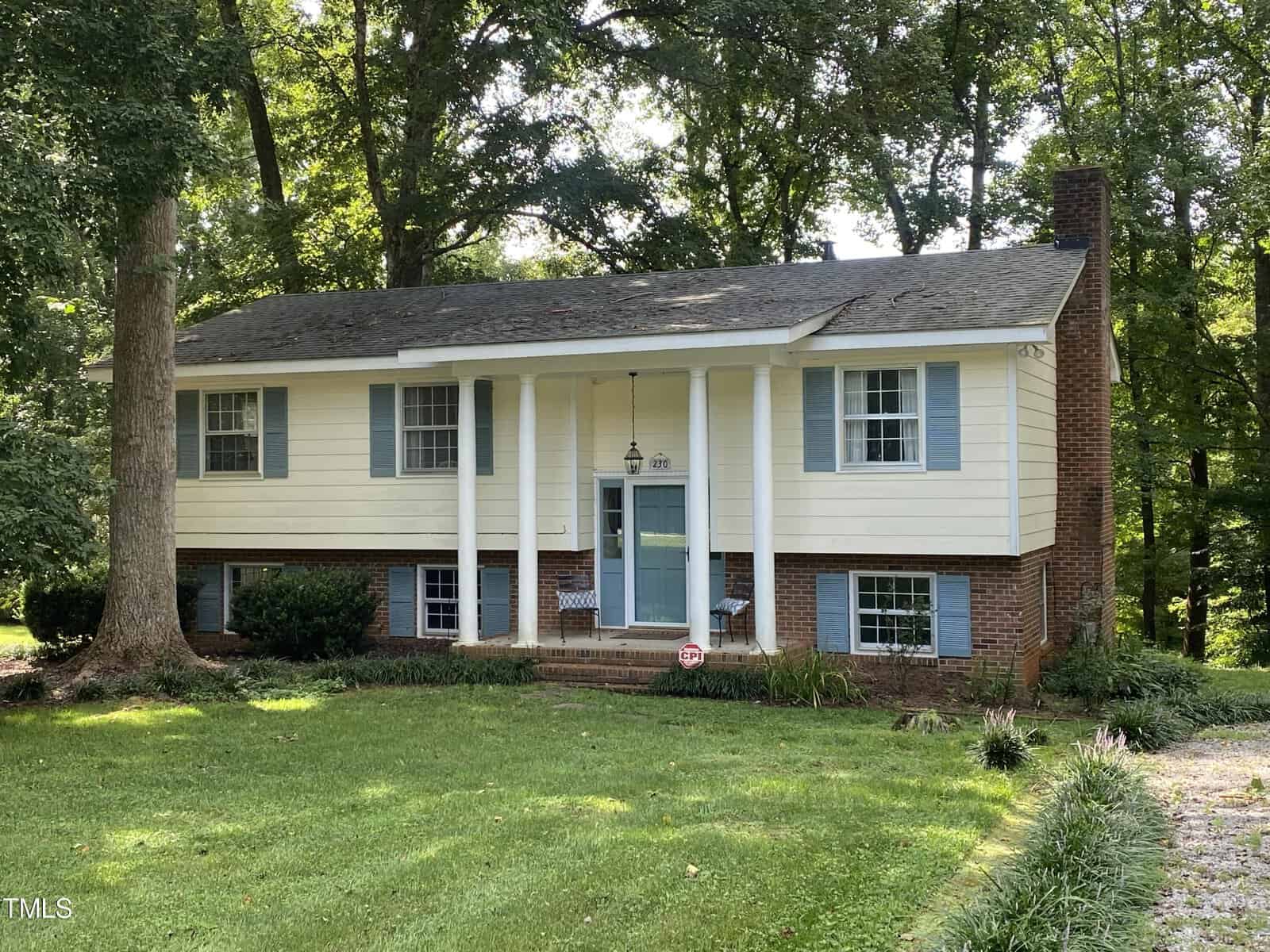 The charming two-story house at 230 Weybossett Road, Henderson, is nestled in a lush, wooded area. The upper portion is painted beige with blue shutters, while the lower half features red brick. The entrance boasts four white columns and a small porch with a hanging light. A well-kept lawn and shrubs adorn the front yard.