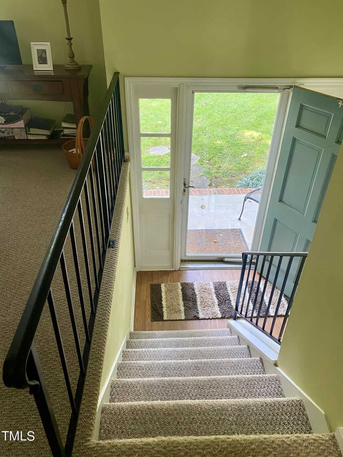 View from the top of a staircase with a light gray carpet, black metal railing, leading to a glass door with a blue frame that opens to a backyard lawn. A striped welcome mat is placed at the bottom of the stairs. A wooden cabinet and basket are visible on the left.