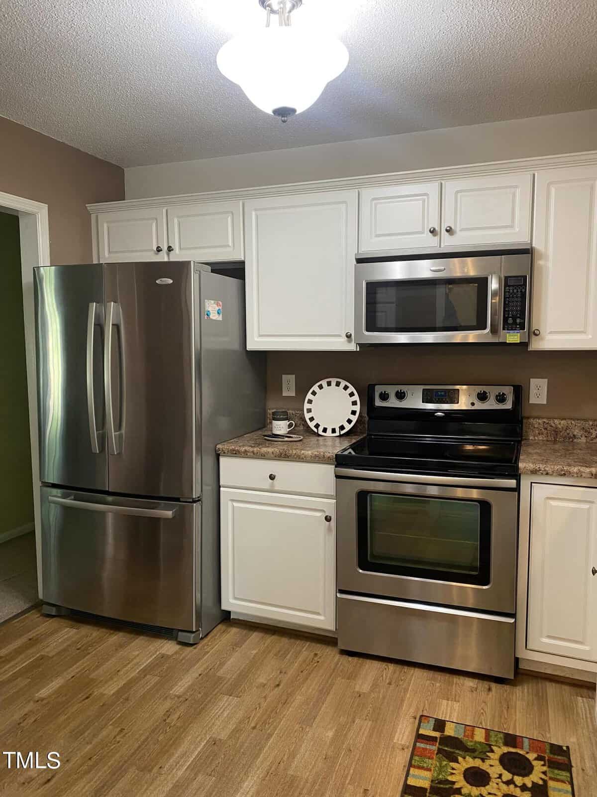 A modern kitchen with stainless steel appliances, including a double-door refrigerator and a stove with an overhead microwave. White cabinets, wooden flooring, and a brown and green patterned rug add to the decor. A decorative plate sits on the counter.