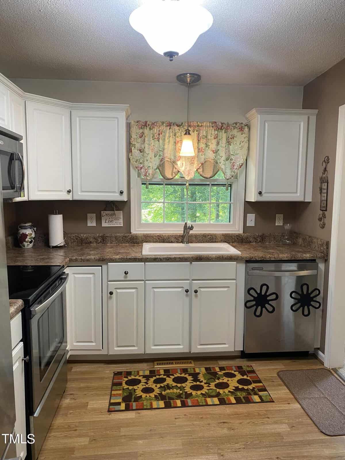 A cozy kitchen featuring white cabinetry and granite countertops. A window above the sink has a floral curtain and an overhead light. Stainless steel appliances, including an oven and dishwasher, are present. A decorative rug and wall decor add charm.