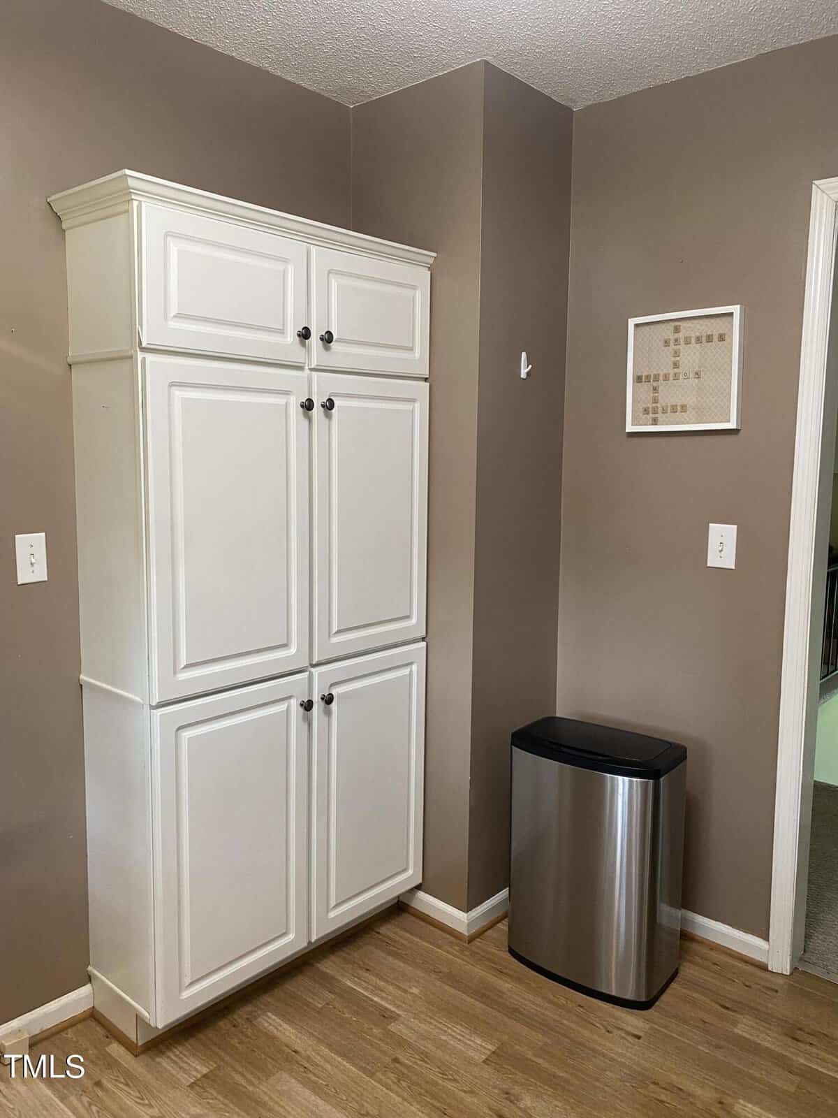 A corner of a kitchen with tan walls and wooden flooring. There's a tall white cabinet with double doors. Next to it is a stainless steel, rectangular trash can. A small framed piece of wall art is hung above the trash can.