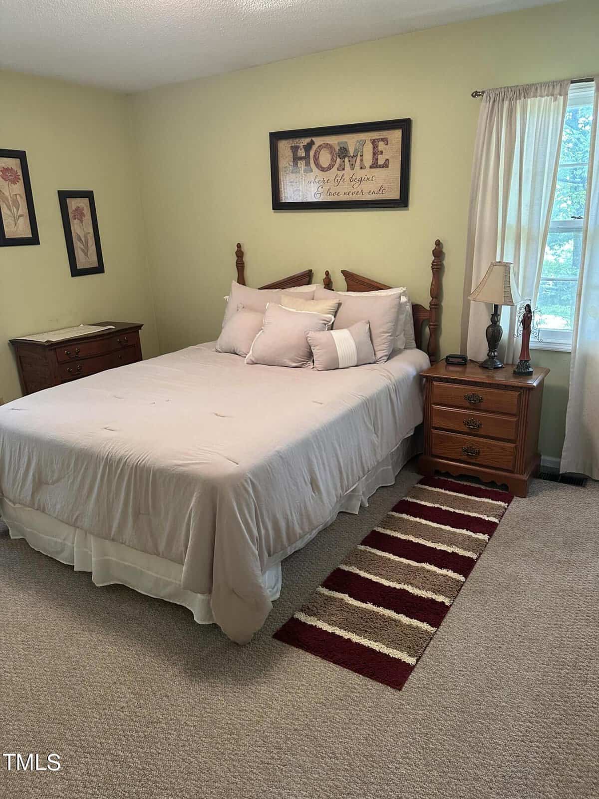 A cozy bedroom featuring a wooden bed with light bedding and pillows. A small wooden nightstand with a lamp sits to the right. Two floral pictures hang to the left, and a framed "HOME" sign is above the bed. A striped rug is on the carpeted floor.