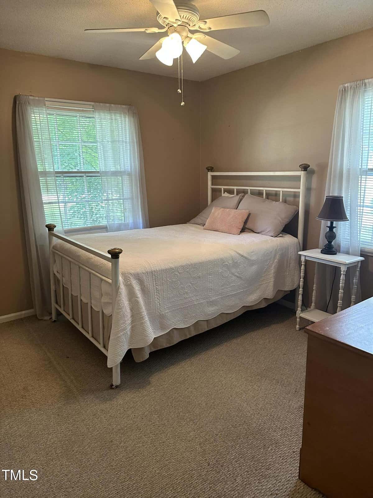 Bright bedroom with a white metal bedframe and white bedding, featuring two pillows. Two windows with sheer curtains let in natural light. A small white bedside table with a black lamp stands next to the bed. The room is carpeted and has a ceiling fan.