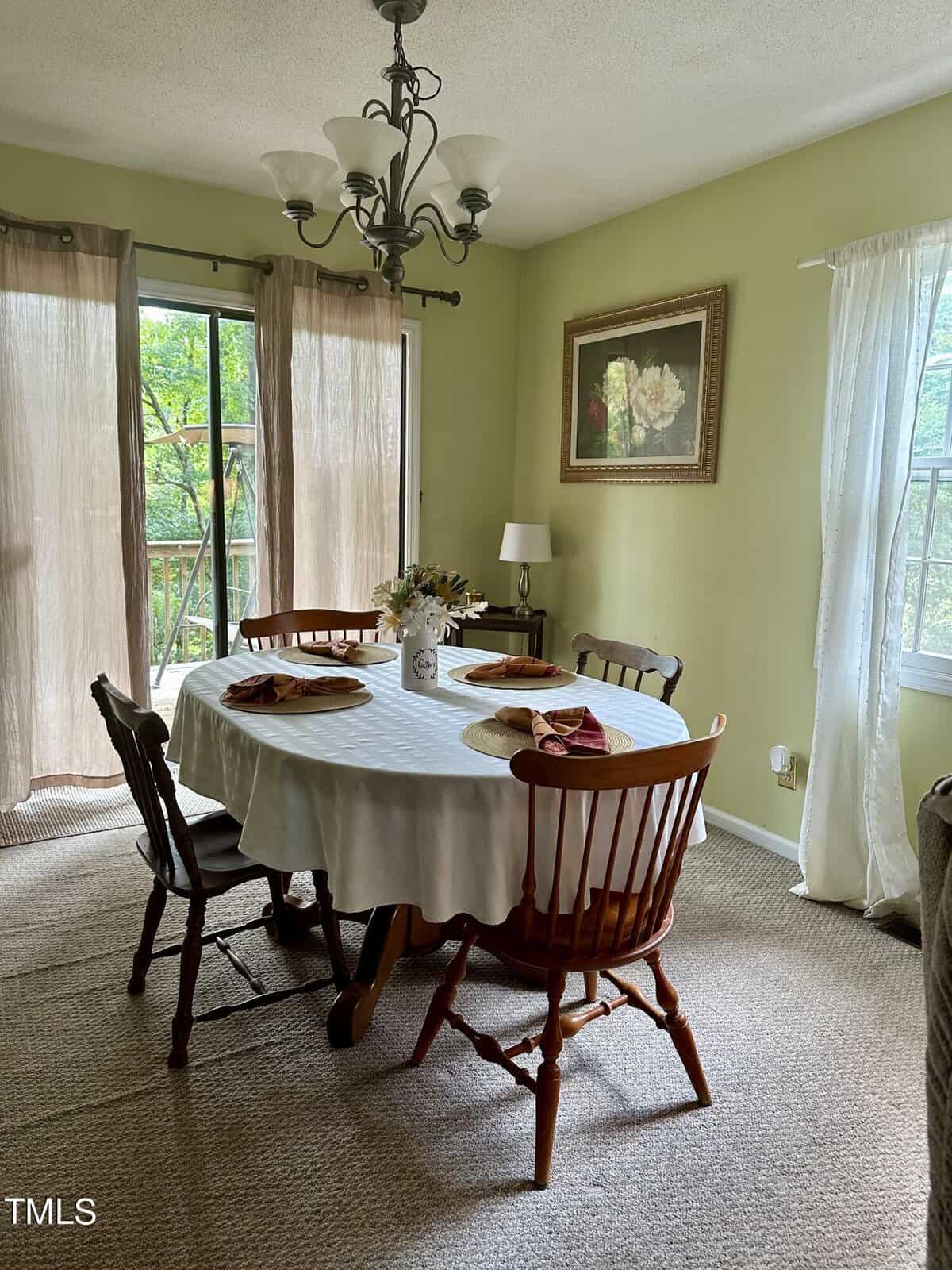 A cozy dining room with a table set for four, draped in a white tablecloth and adorned with a centerpiece of flowers. There are four wooden chairs around the table. The room features light green walls, a hanging chandelier, a framed painting, and large windows with sheer curtains.