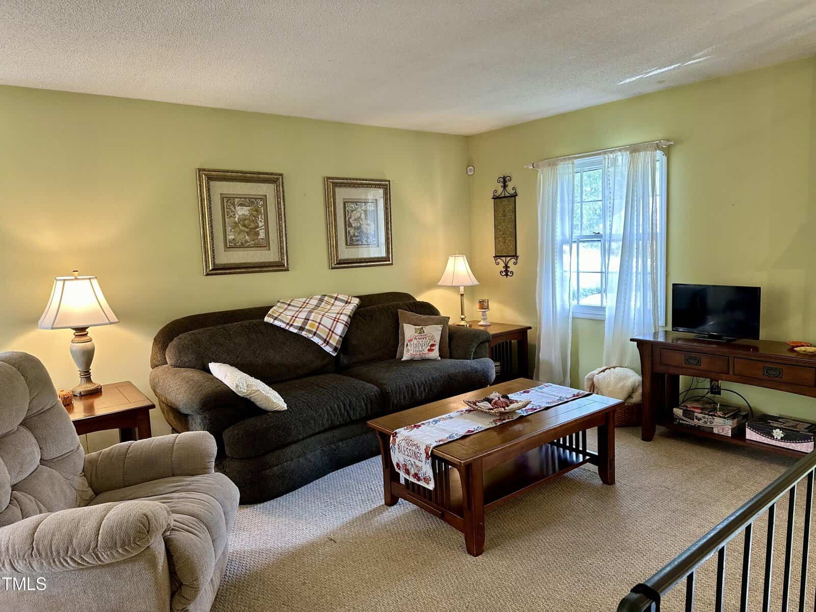 A cozy living room with pastel green walls, furnished with a brown sofa, a beige recliner, and a wooden coffee table. Two framed pictures adorn the wall above the sofa. A small flat-screen TV sits on a wooden stand near a window with white curtains, allowing natural light to enter.