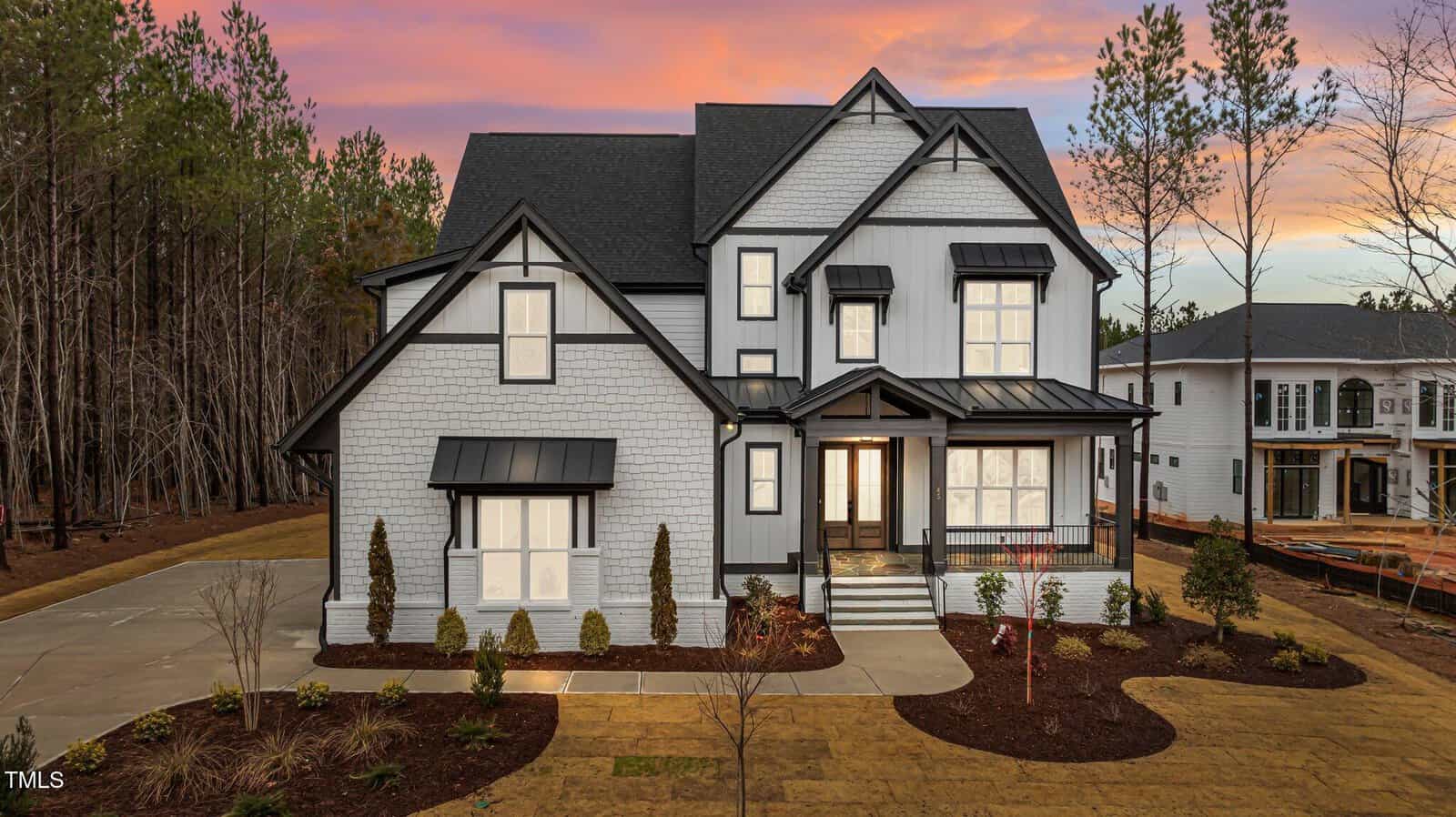 Two-story modern farmhouse with white siding, black trim, large windows, and a covered front porch. The home is surrounded by newly landscaped yard and tall trees, with a vibrant sunset sky in the background.