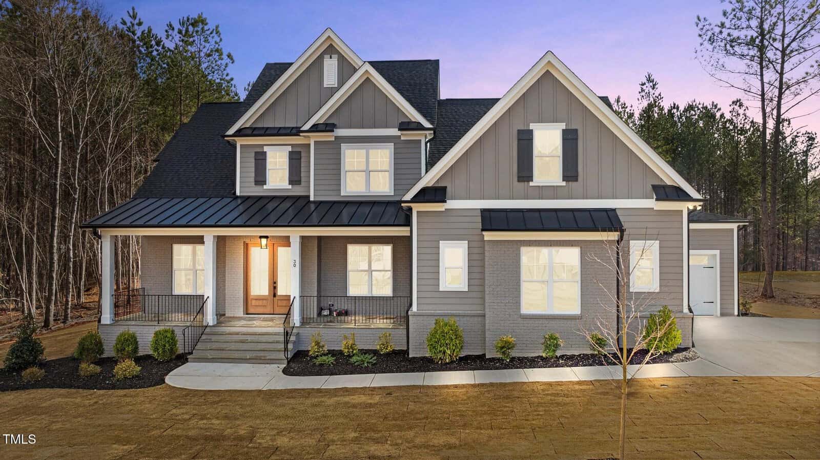 A modern two-story house with gray siding, white trim, black shutters, and a covered front porch with railings, set against a backdrop of trees at dusk. A concrete driveway leads to a garage on the right.