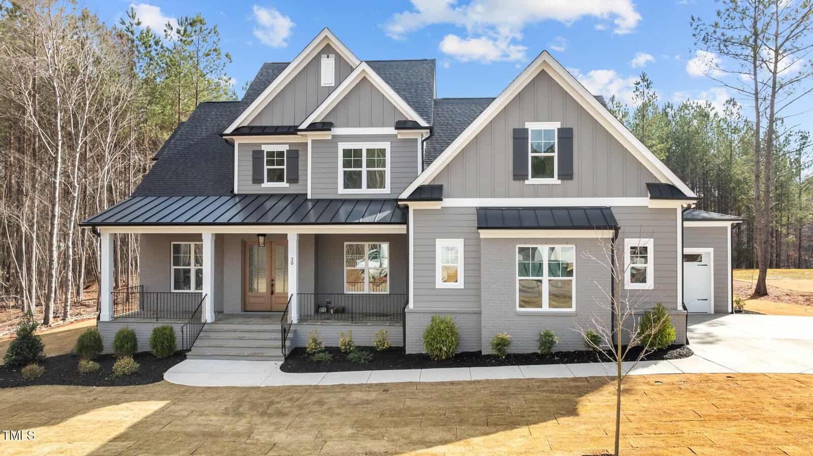 A modern two-story gray house with white trim, black shutters, and a covered front porch. The home features large windows, a gabled roof, and a driveway leading to a side garage, surrounded by a neat lawn and trees.