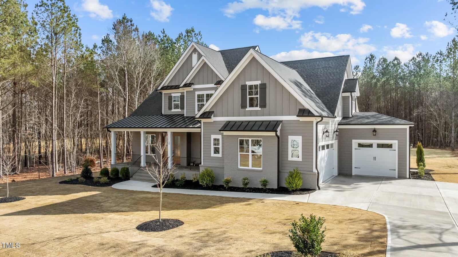 A modern two-story gray house with white trim, front porch, attached garage, and landscaped yard, set against a backdrop of trees and blue sky.