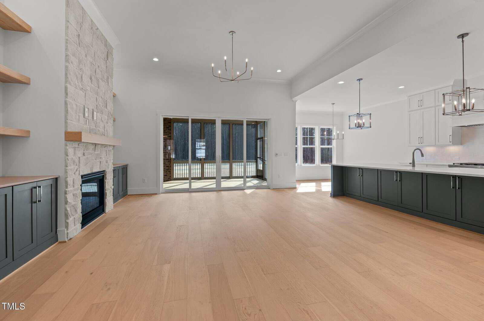 A spacious, modern open-concept living area with light wood floors, a stone fireplace, built-in shelves, large kitchen with white cabinets, pendant lighting, and sliding glass doors opening to an outdoor patio.