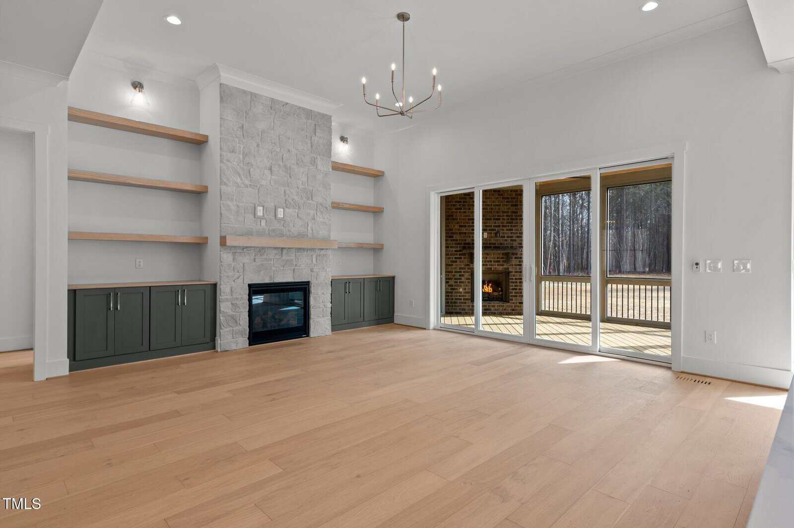 A modern, empty living room with light wood floors, a stone fireplace, built-in shelves and cabinets, and large glass doors opening to a deck with a view of trees and a brick outdoor fireplace.