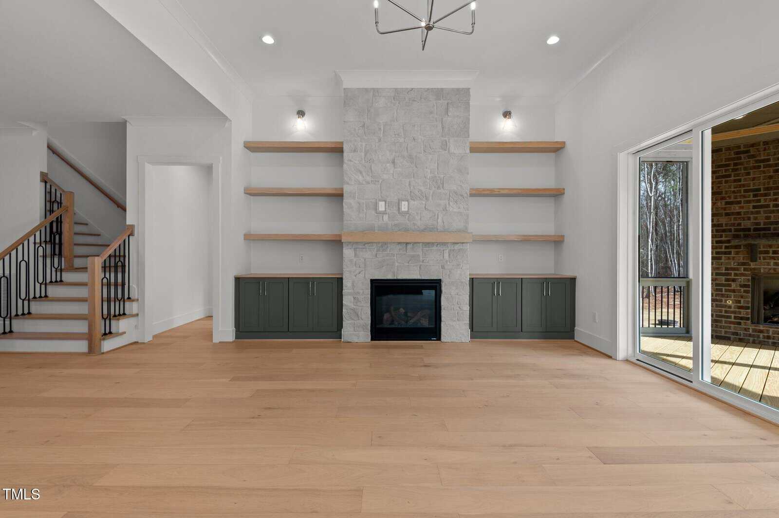 Modern living room with light wood floors, a stone fireplace, built-in shelves and cabinets, recessed lights, view of staircase on the left, and sliding glass doors opening to a covered patio on the right.