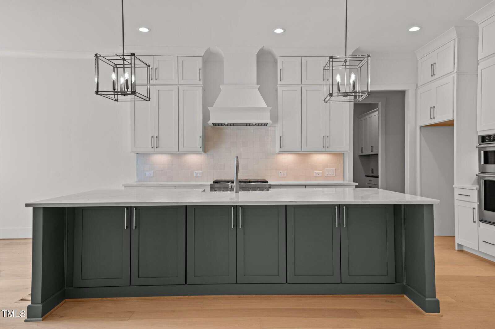Modern kitchen with white upper cabinets, a large gray island with a sink, stainless steel appliances, light wood flooring, and two geometric pendant lights hanging over the island.