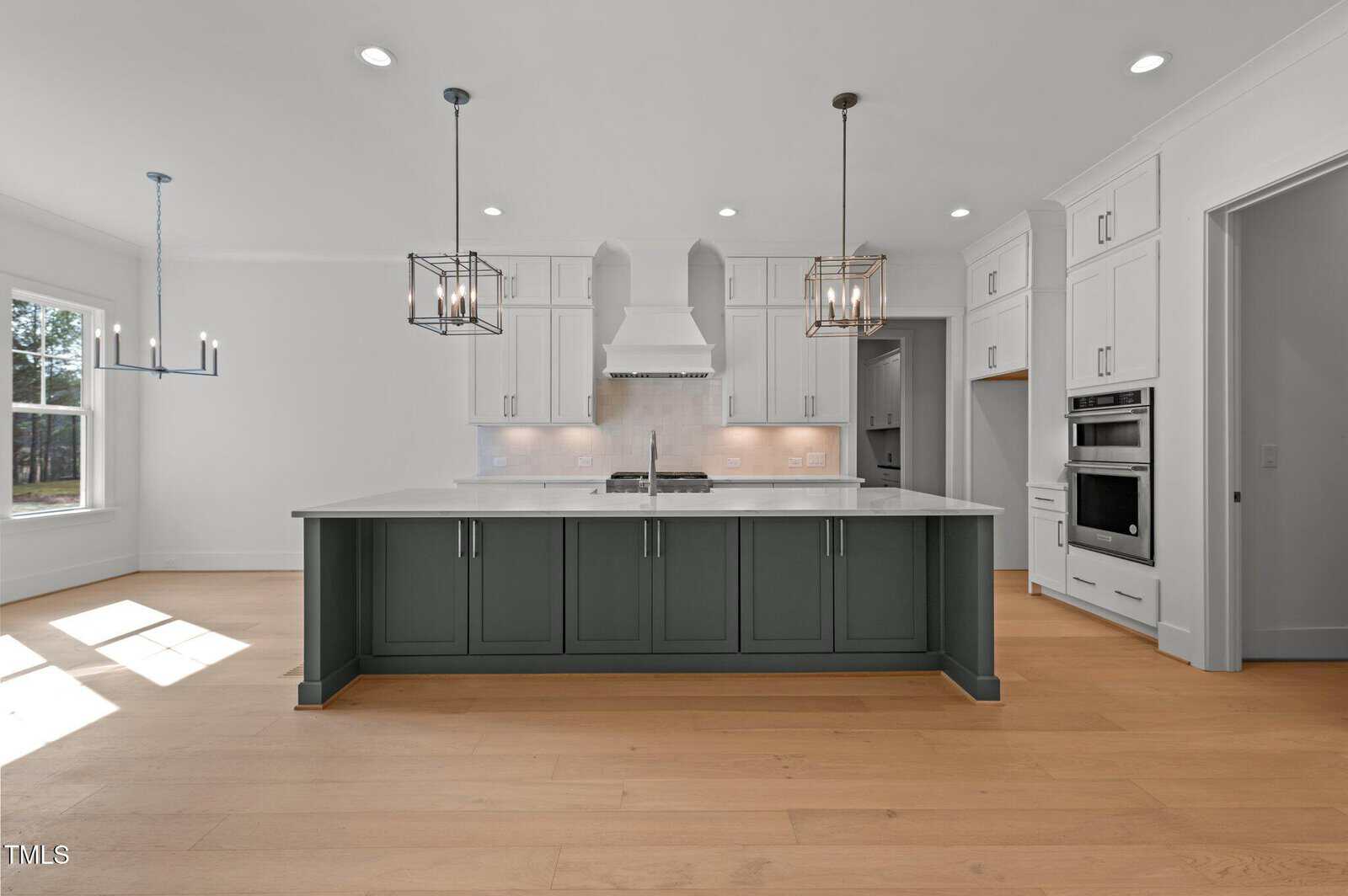 Modern kitchen with a large island featuring dark gray cabinets, white countertops, pendant lights, white shaker cabinets, built-in oven, range hood, and light wood flooring, with natural light from windows on the left.
