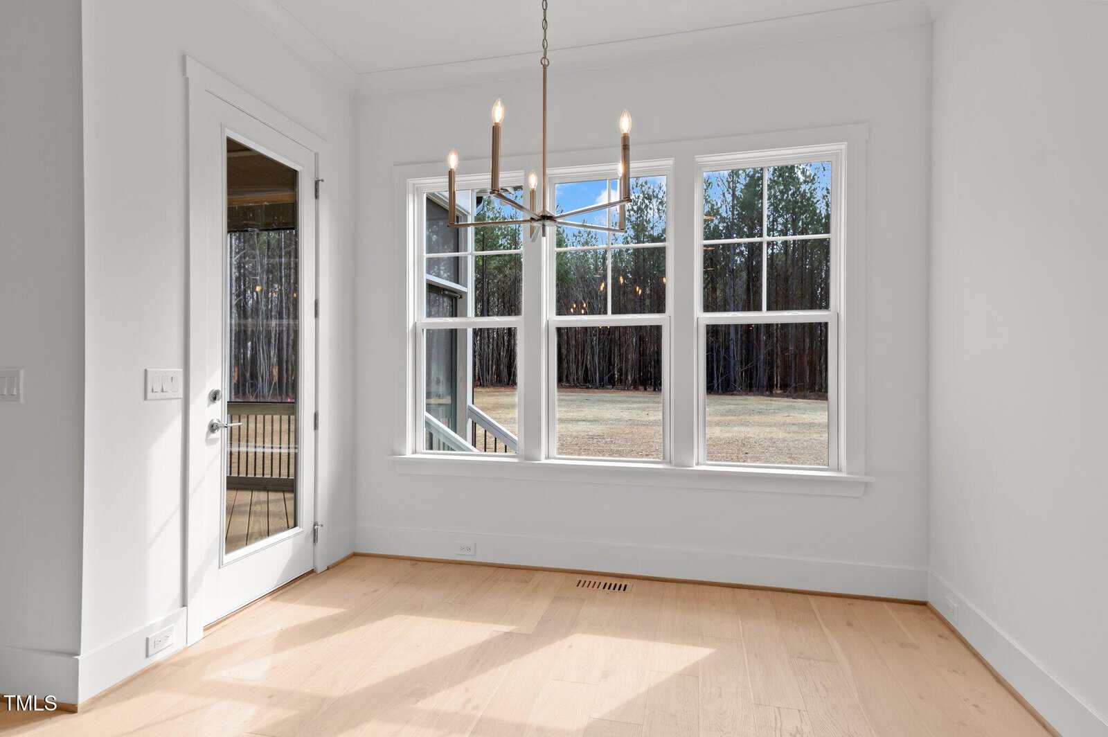 A bright, empty room with light wooden floors, large windows letting in natural light, a modern chandelier, and a glass door leading outside. A wooded area is visible through the windows.