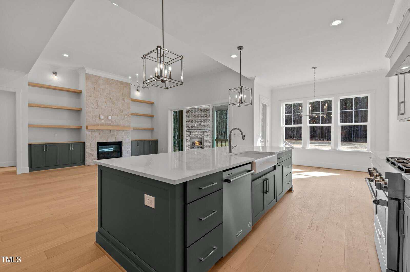 Modern kitchen with a large island, dark cabinets, white countertops, pendant lights, and stainless steel appliances. An open living area with built-in shelves, a stone fireplace, and large windows letting in natural light.