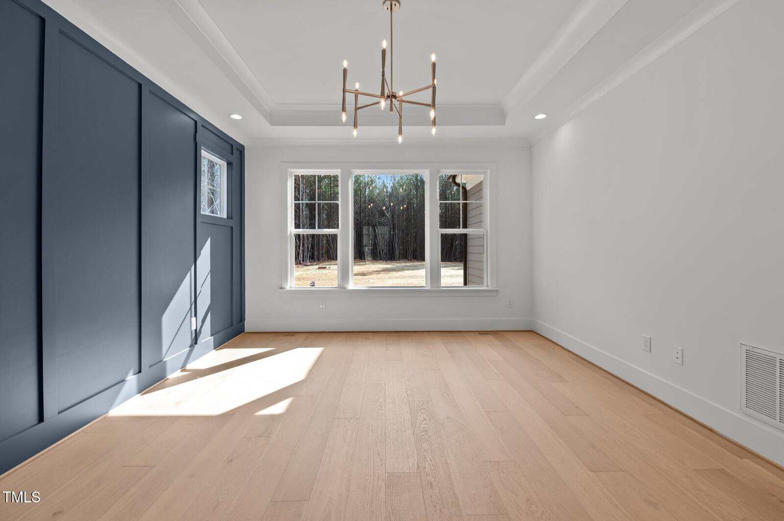 Bright, empty room with light wood floors, blue accent wall, modern chandelier, and large windows letting in natural light.