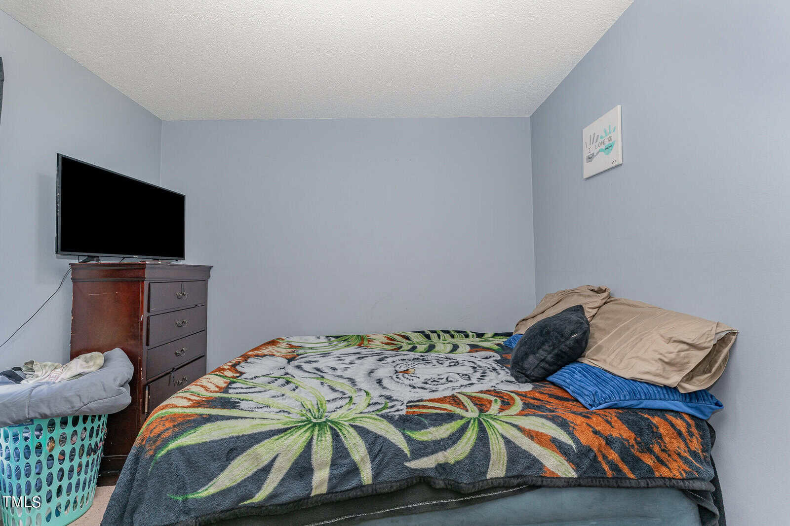 A bedroom with light blue walls features a bed covered in a colorful blanket with a tiger design. There's a stack of pillows and a flat-screen TV on a brown dresser. A laundry basket is nearby, and a small wall decoration is visible.