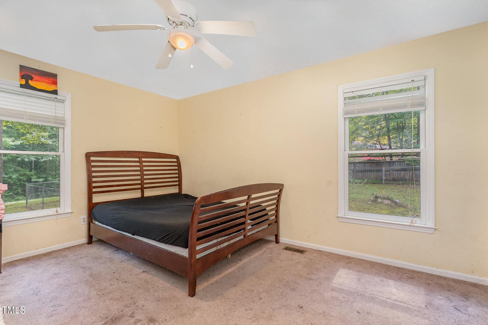 A sparsely furnished bedroom with beige walls featuring a large wooden bed with a black sheet. Two windows provide natural light, revealing a view of greenery outside. A ceiling fan hangs above, and a small piece of art adorns the wall.