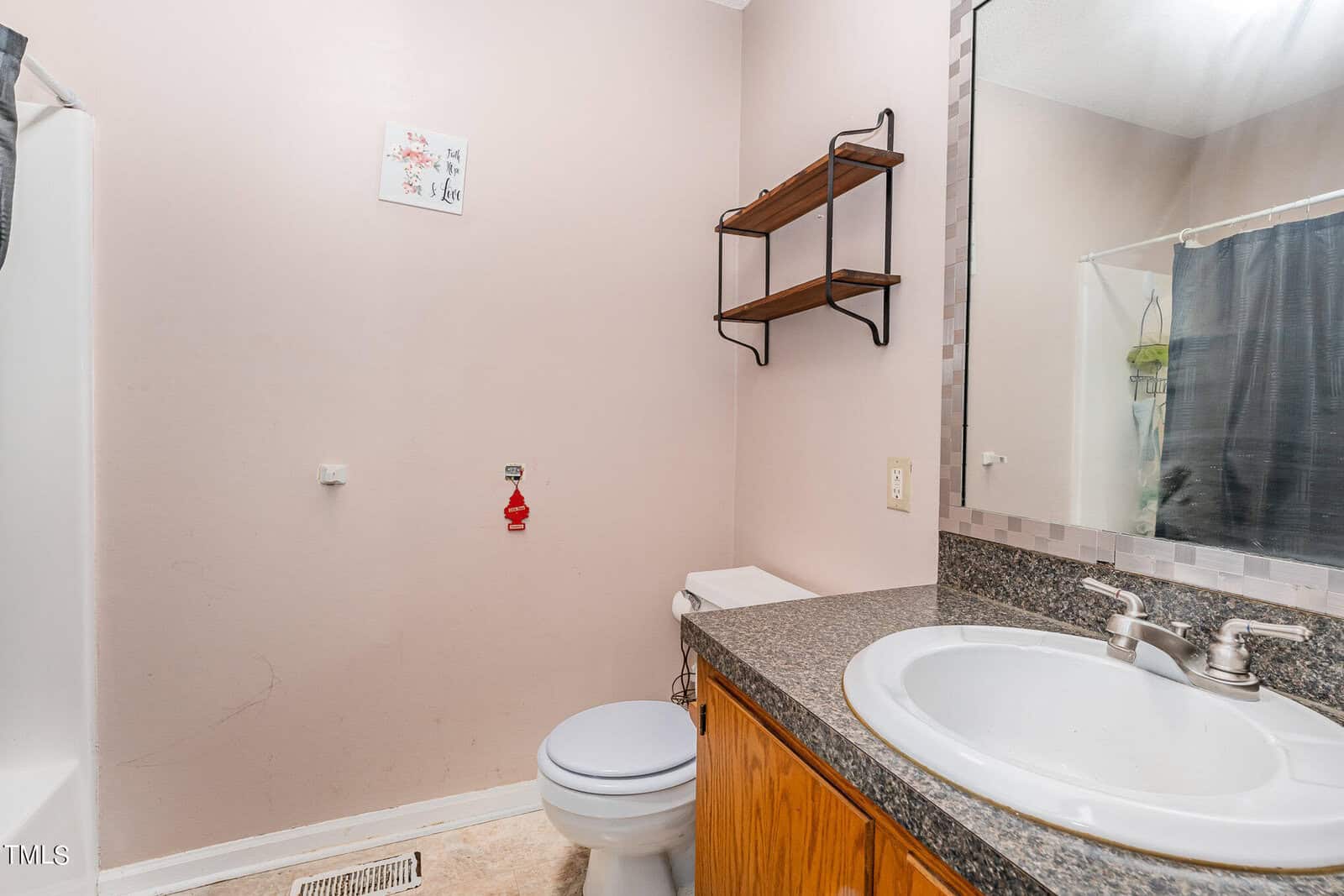 A small bathroom with a granite countertop, white sink, and wooden cabinets. There's a toilet next to an empty wall space for storage. Above, a set of shelves is mounted. A shower is partially visible with a curtain partially open.