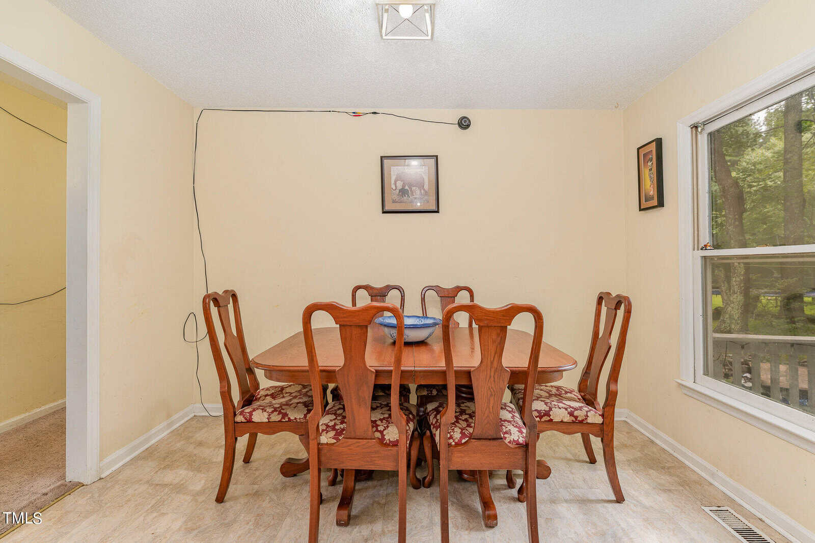 A dining room with a wooden table and six matching chairs, each with floral-patterned cushions. A framed picture hangs on the wall above the table, and a window to the right shows trees outside. A small dish is centered on the table.