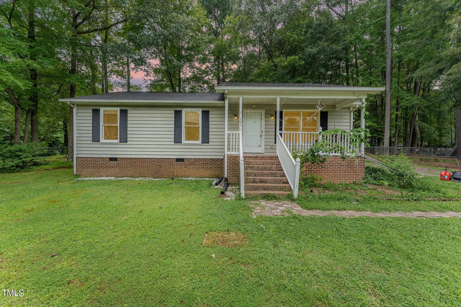 A small, single-story house at 215 E Quailridge Road features light siding and dark shutters, nestled amidst lush green grass and tall trees in Oxford. The charming home boasts a front porch with steps leading up to it, set against the serene backdrop of dense woods.