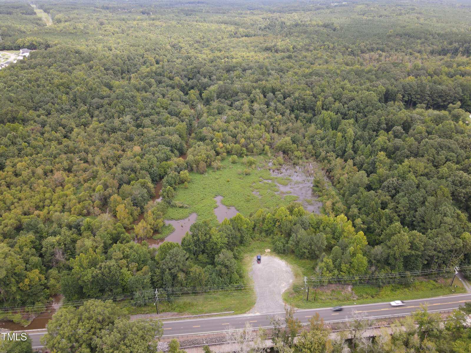 Aerial view of a dense, green forest with a small clearing near Creedmoor. A road, 2503 NC Hwy 56, runs below the forest, leading to a parking area with one car. Lush trees and greenery stretch into the distance under an overcast sky.