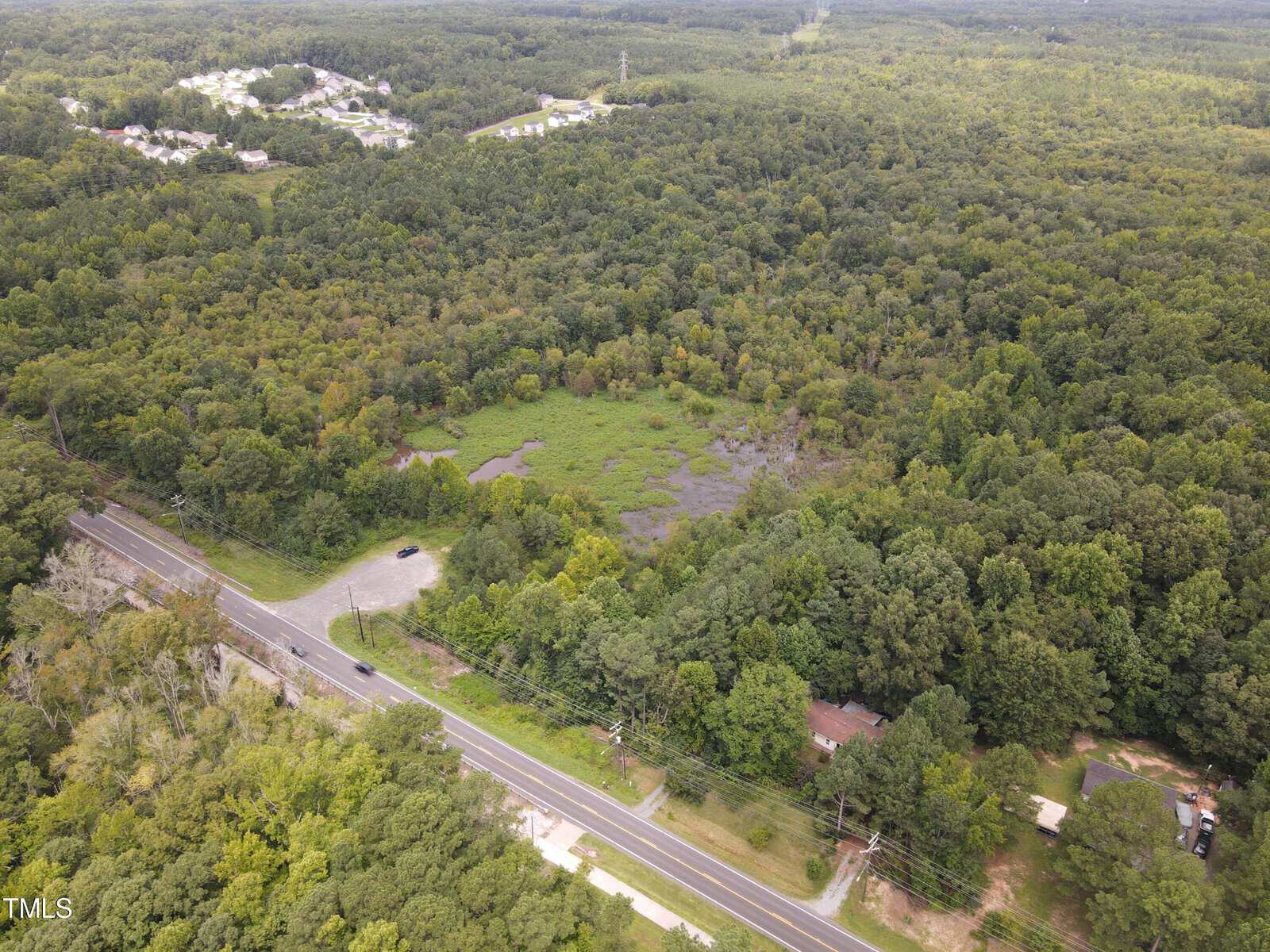 Aerial view of a lush green forest with a road cutting through the bottom left corner. There are scattered houses and a neighborhood visible in the background, set amidst dense tree cover and patches of open land.