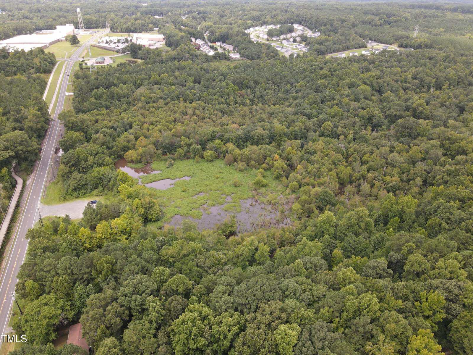 Aerial view of a lush, green forest surrounding a small clearing with a pond. Roads border the forest on the left, with industrial and residential areas visible in the background under a cloudy sky.