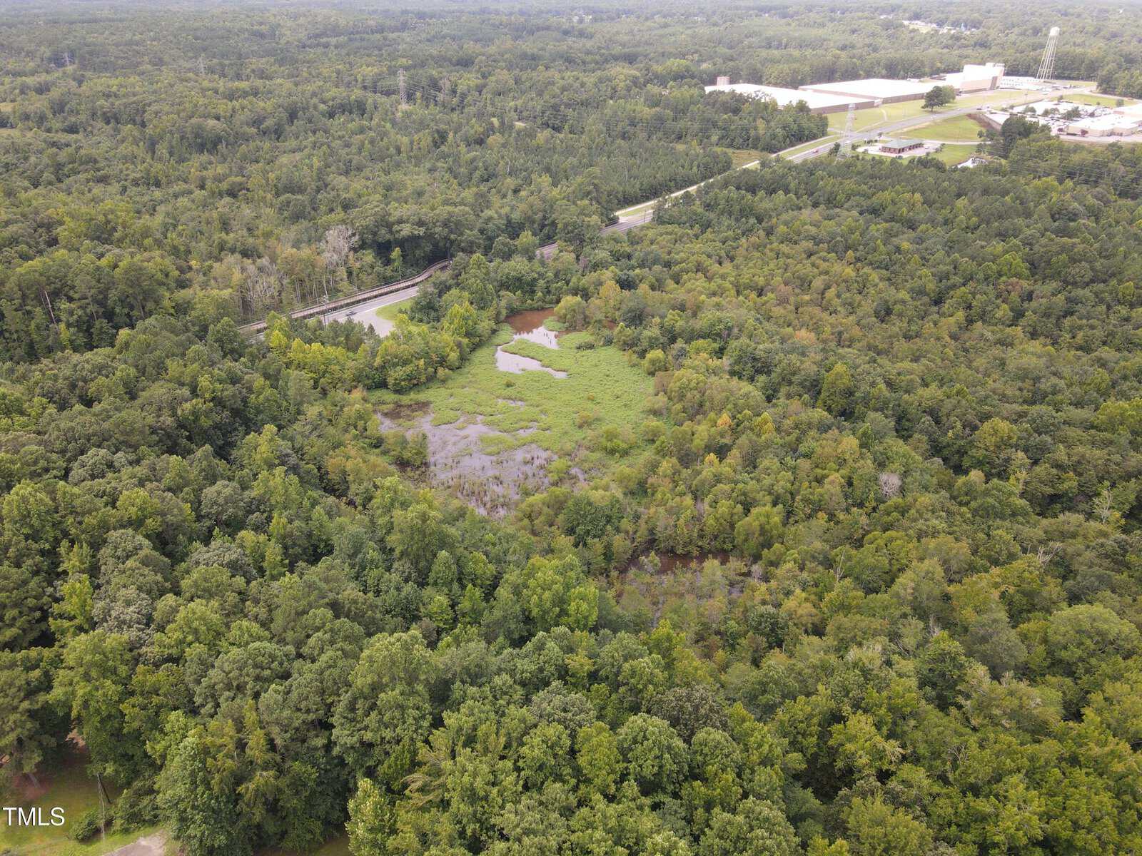 Aerial view of a dense green forest with a small wetland area in the center. Beyond the forest, there are roads and a few industrial buildings. The landscape stretches into the horizon under a cloudy sky.